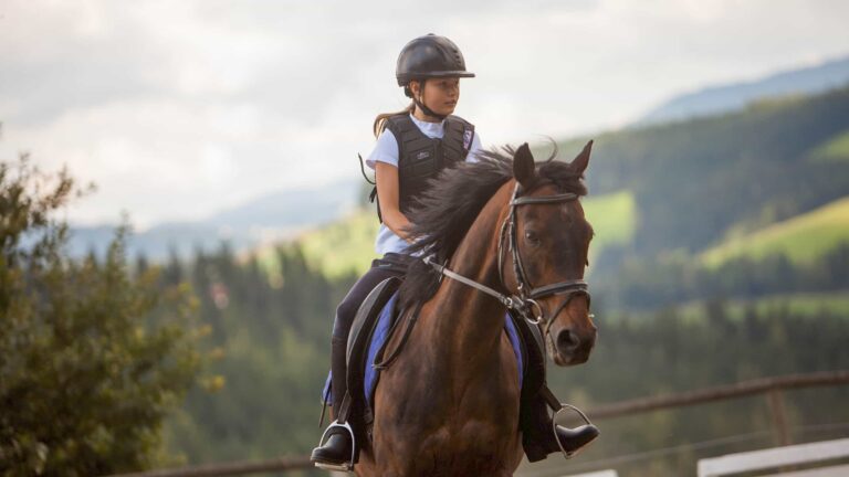 Familienhotel Der Ponyhof, Familotel Steiermark: junges Mädchenm reitet braunes Pferd auf Reitplatz