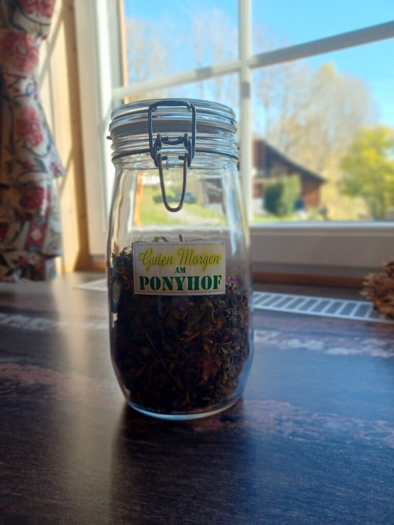 Glas mit Kräutertee und dem Etikett „Guten Morgen am Ponyhof“ auf einem Holztisch vor einem Fenster mit Blick ins Grüne.